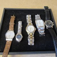 Five assorted watches laid side by side on black background showing variety of styles and bands