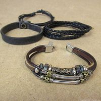 Three vintage style bracelets displayed on a beige surface: one wide black leather strap, one thin multi-strand black braided leather, and a brown leather bracelet with metal beads and tubular accents.