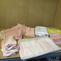 Full view of the assortment of folded hand-knit doilies and textiles laid out on table showing various fabrics and patterns, mostly pastel pink and white.