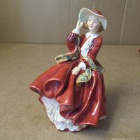 Front and side view of the Royal Doulton 'Top of the Hill' figurine showing seated woman in red dress and white wide-brimmed hat with red band, detailed multicolor sash over shoulder.