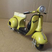 Front side view of the yellow Vespa scooter figurine showing the Vespa logo on the front panel, headlight, and handlebars.