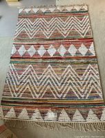 Full view of woven area rug showing geometric patterns with zigzags, diamonds, and triangles in white over a multicolored striped background. Fringe visible on the shorter edge.
