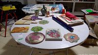 Full table showing assortment of decorative art plates, cutting boards, vases, copper wine cups, and art supplies