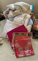 Pile of assorted linens including pillows, comforter, cushions, pillowcases, and saris in folded stacks showing fabric textures and colors.