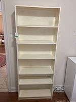 Front view of white laminate bookshelf with five shelves, clean surface, and cable cutout in back panel near bottom.