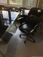 Black office chair next to glass office desk with plastic floor mat underneath it, setting shows office environment.