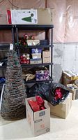 View of shelving with boxes of Christmas ornaments, wrapped gifts, faux poinsettias, and gift wrap supplies, with the Ashland 7ft pre-lit Christmas tree box visible on top shelf and large wood Christmas tree in front