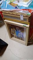 Stack of various wooden picture frames including one white distressed frame and one ornate gold frame with prints visible inside.
