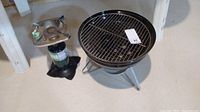 Photo showing propane camping stove and Weber tabletop charcoal grill side by side on floor.