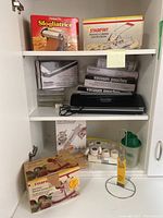 Photo of white cabinet shelving with assorted boxed kitchen items: Imperia pasta maker, Starfrit potato chipper, Sous Vide Supreme vacuum sealer and vacuum pouches, cake decorating set, salad dressing mixer, paper towel holder.