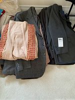 Pile of men's wool jackets and suit pants stacked on the floor with a folded beige kaftan on top, showing texture and button details of jackets.