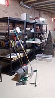 View of four black plastic shelving units in basement, with various items stored on and around them. Shelves have grid-style surfaces and are five-tiered, approx. 35x24x73 inches each.