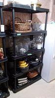 Two black resin shelving units side by side in basement setting with various household and kitchen items stored on each shelf