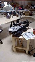 Wood table with scroll saw and attached white lamp in basement area with other items in background