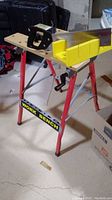 Full view of the wood and metal work bench with a yellow mitre box and hand saw positioned on top, showing red steel legs and wooden surface.