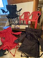 Photo of three folding bag chairs (blue, red, black), two red kids plastic chairs and the dome tent in original packaging arranged on a wooden surface