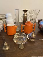 Close-up of glass vase, orange ceramic pots, brass vase, glass ashtray, and brass bell on wooden surface.