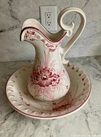 Beige ceramic wash basin and pitcher with red floral decoration placed on a stone surface.