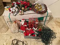Lot of Christmas items arranged on and around an artificial Christmas tree box; includes Santa figurine, angel figure, red baubles, floral stems, decorative wreath, string lights, and other decorations.