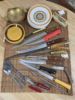 Various cutting utensils including kitchen knives, pocket knives, skewers, corkscrew, a spoon, vintage plates, and a cooper ashtray arranged on a mat.