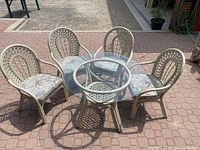 Overview of lot showing round glass top table with beige wicker base and four matching beige wicker chairs with floral seat cushions arranged around table on outdoor brick pavement.