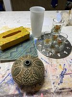 All items shown together on table: oval Nicaraguan pottery, yellow tissue box, white vase, Alessi tray with five skull design glasses