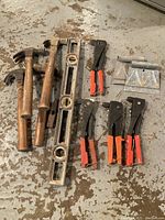 Lot of used hand tools including three wooden-handled claw hammers, one spirit level, four pop rivet guns with orange handles, and three metal carpenter squares placed on floor.