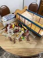Full view of wooden doll bed with floral bedding, soft doll in patterned clothes, and assorted plastic animal toys scattered on table.