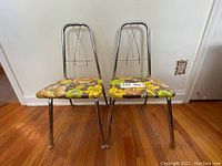 Pair of vintage kids chairs with chrome tubular frames and floral vinyl covered seats, positioned side-by-side on wooden floor against wall.