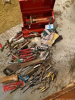 Photo showing open red metal toolbox with hammers inside, surrounded by various hand tools including wrenches, pliers, gloves, and safety glasses on the floor.