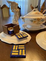 Photo showing blue and yellow tile coasters in wooden box with soup tureen and vase in background.