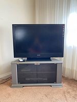 Sony flat-screen TV on top of grey TV stand with glass doors placed on carpeted floor near window.