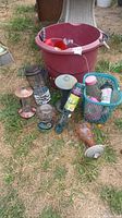 Photo showing assortment of multiple bird feeders and a large red bucket with feeders and parts inside, placed on grass and dirt surface.