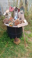 Collection of assorted birdhouses placed on a black barrel outdoors surrounded by grass and weeds
