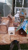 Lot of various birdhouses displayed on plywood surface outdoors. Includes natural wood, painted, and license plate-roof styles. Weathered condition visible.