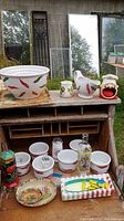Overview photo of the full lot showing large mixing bowl with chili design, soup cups, pitchers, decorative bottles, corn cob trays with skewers, sponge holder shaped as a chef, small salt shaker and other kitchen items on a wooden desk outdoors.