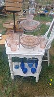 Pink and blue glass dishware arranged on small white table outside, showing pink glass cups, plates, bowls, and a two-tier cake stand on top shelf; blue glass bottles and bowls on lower shelf.