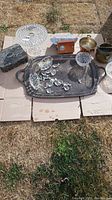 Full lot on cardboard outdoors, showing crystal cake plate, small radio, tin box, glass bowl, and metal serving tray with additional small glass items on top.