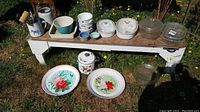 Wide shot of all items displayed on a wooden bench outdoors in grass, showing flowered pot with lid, two large floral glass platters, white ceramic canisters, two clear glass Pyrex baking dishes, and four bowls stacked.
