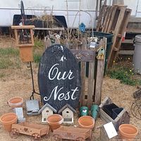 Wide view showing all items: 7 terracotta flower pots, various small wooden birdhouses, two rectangular metal bird feeders, a wooden wall sign reading 'Our Nest', and a tall wooden planter resembling a picket fence.