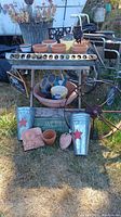 Wide shot of gardening lot showing metal table with terracotta pots, galvanized metal buckets, antique metal wheel, wooden garden signs, and terracotta wall hanging on grass outdoors.