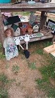 Photo of assorted garden decorations including two wooden birdhouses with green roofs, decorative painted wooden figures, metal garden stakes with cow designs holding a 'GARDEN' sign, and small flower pots on a wooden shelf outdoors on grass.