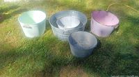 Wide view of all five metal buckets placed on grass showing various sizes and colors including purple, green, galvanized, and white finish with some rust spots.