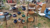 Wide view showing multiple ceramic flower pots in blue, green, and purple on rust-colored stand and black metal stand with coir lined baskets, outdoor setting.
