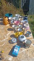 View of entire assortment of about 20 flower pots arrayed on wooden surface outdoors. Various sizes, colors, and materials including ceramic, plastic, and metal.