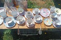 Multiple tea cups and saucers with various floral patterns displayed on a wooden surface outdoors, showing general condition and variety.