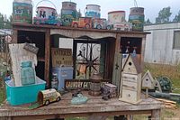 Overview of lot showing various vintage decorative tins with colorful scenes, wooden birdhouses, metal and wood signs, and miniature metal toys on a rustic wooden table outdoors.