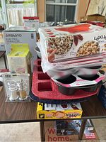 Stack of baking pans including red speckled sandwich pan, muffin pans, Lékué popcorn popper box, Ambiano electric spiralizer box, and salt and pepper shakers set on table.