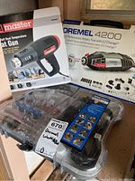Photo of Drill Master 1500 Watt Dual Temperature Heat Gun box, Dremel 4200 rotary tool box, and rotary tool accessory set in a plastic carrying case together on a surface near a window.