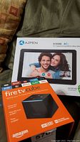 Shows the boxed Azpen tablet and Fire TV Cube box alongside each other on a couch.